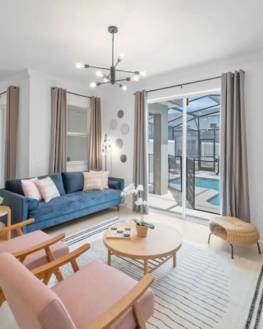 Bright open-plan living room with blue sofa, pink chairs, round wood table, and glass doors opening to pool area